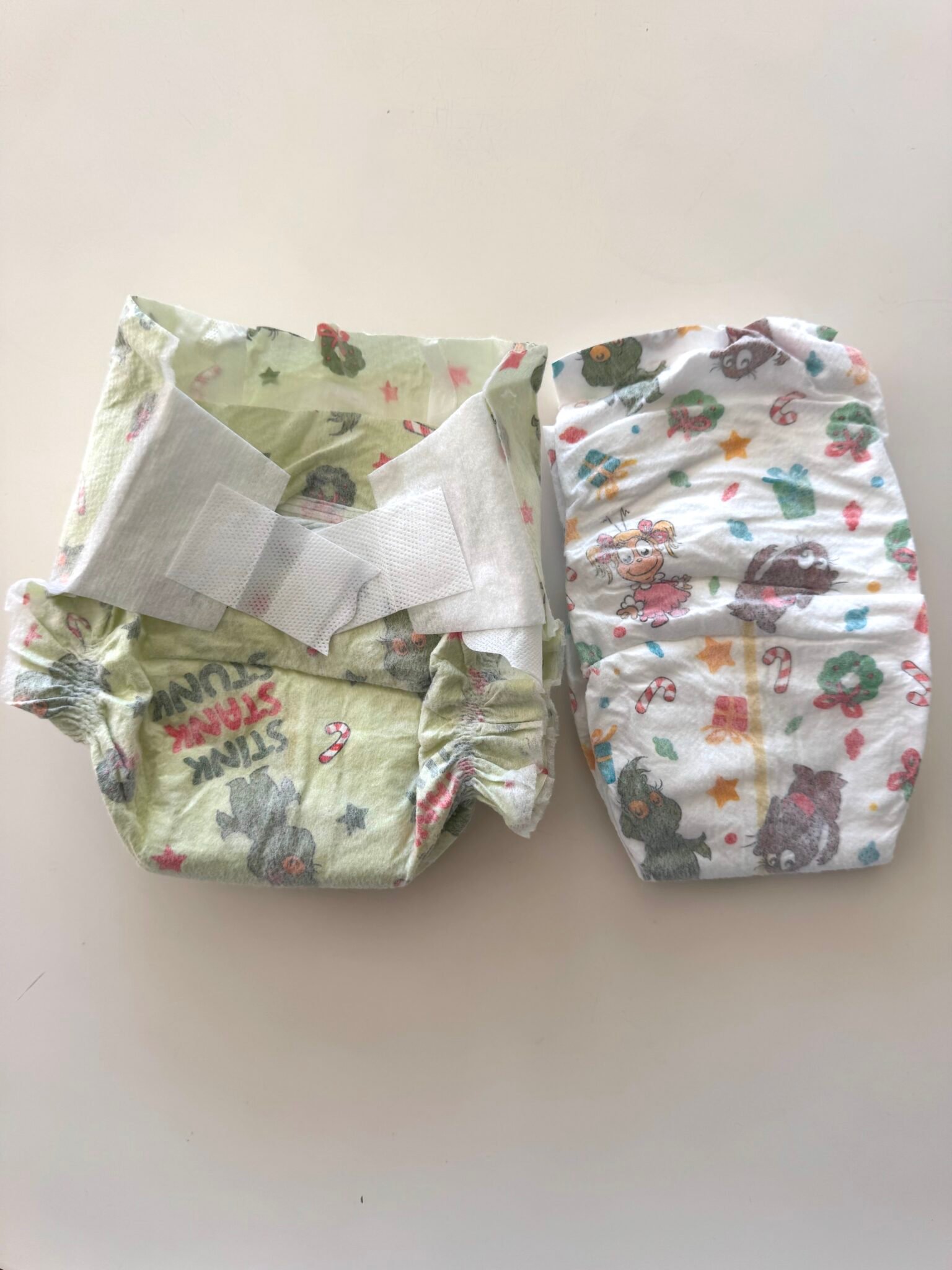 Two disposable baby diapers with cartoon animal prints lie side by side on a white surface; one diaper is open and the other is folded.