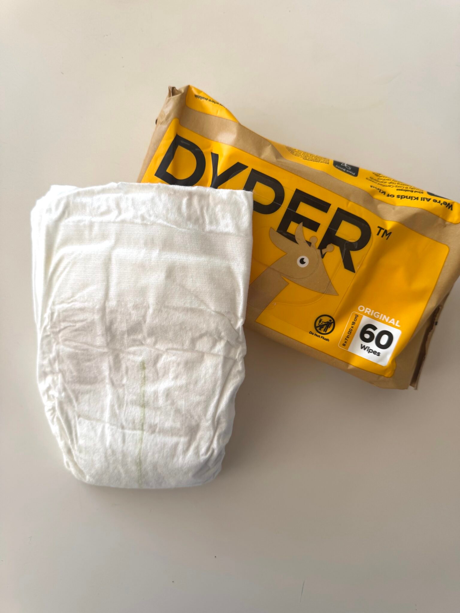 A white disposable diaper and a yellow package labeled "DYPER Original 60 Wipes" are placed on a white surface.