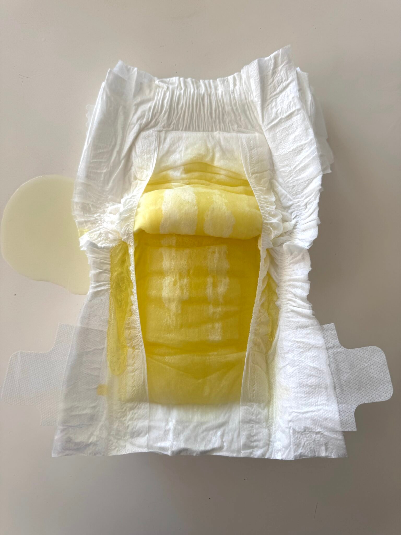 A disposable diaper with yellow liquid visibly soaked into the absorbent area and leaking slightly onto a white surface.
