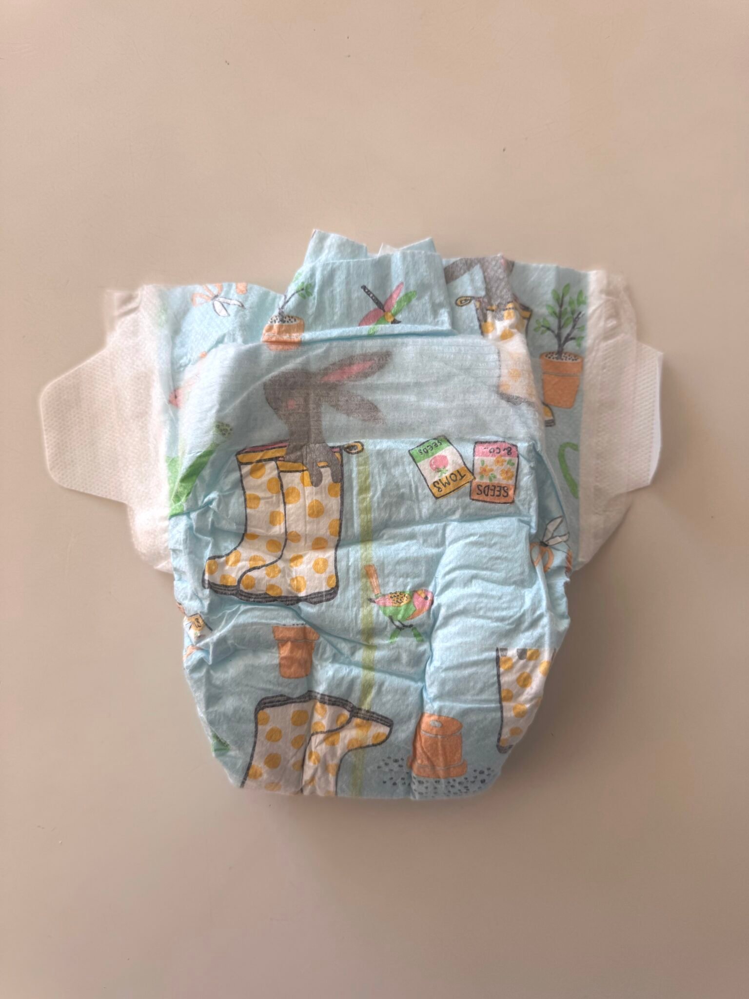 A disposable baby diaper with colorful printed designs, including yellow rain boots, plants in pots, and seed packets, lying on a plain surface.