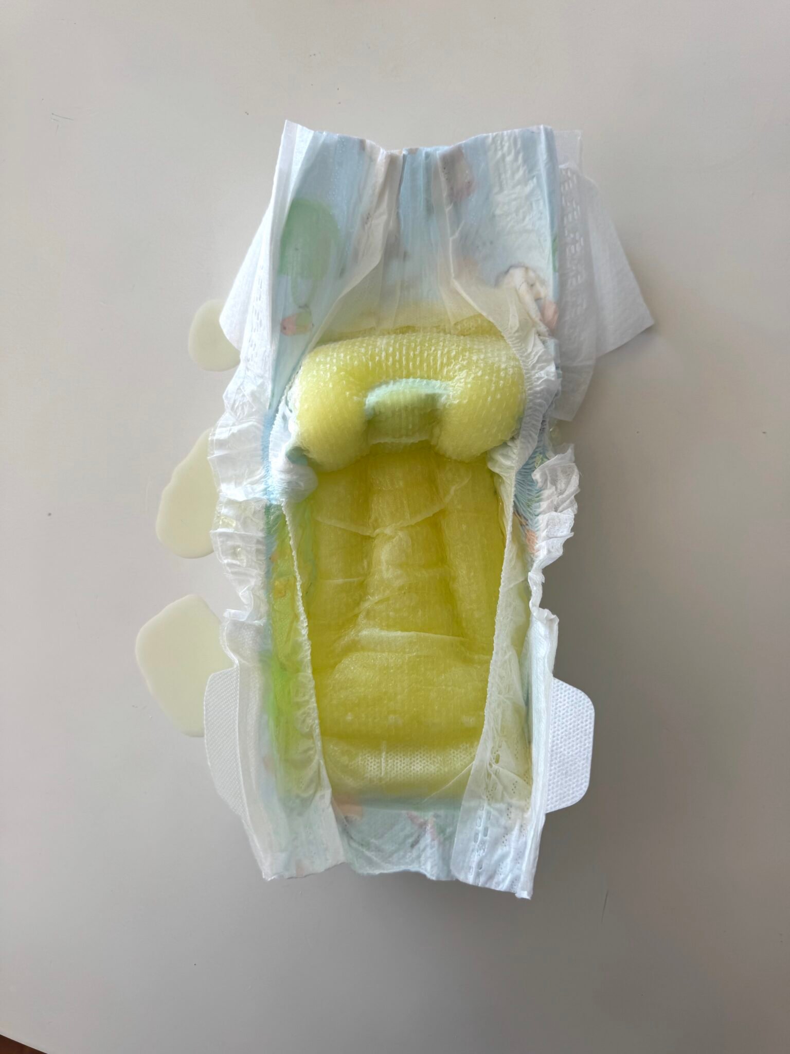 An opened disposable diaper with yellow liquid absorbed inside, showing leakage on the surrounding white surface.