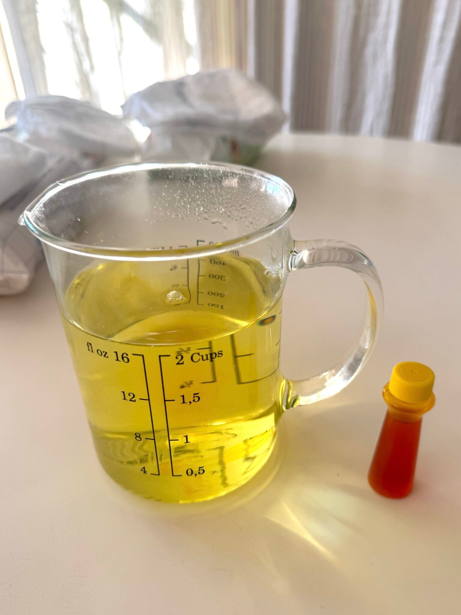 A glass measuring cup with yellow liquid sits on a white surface next to a small plastic bottle with a yellow cap.