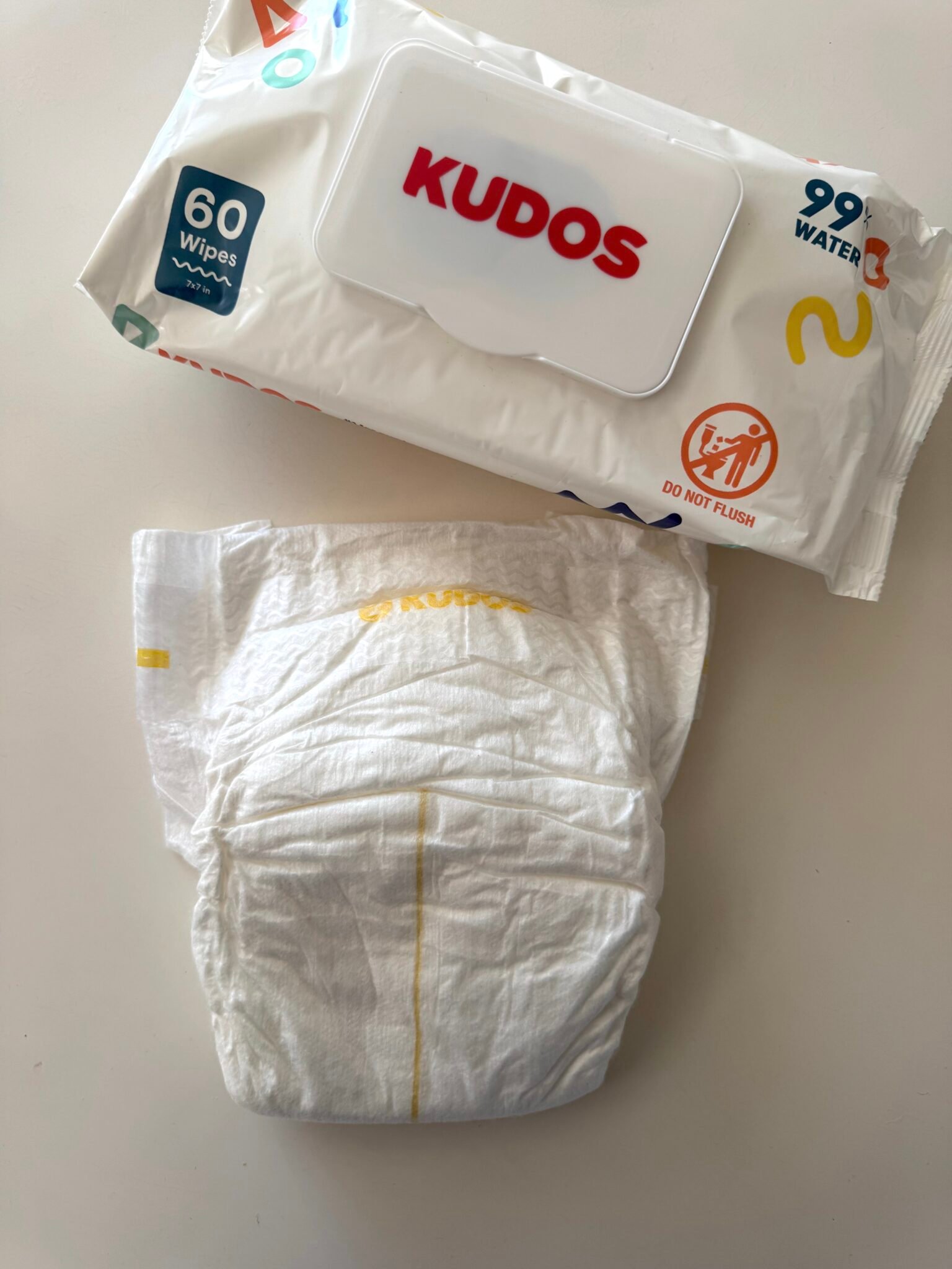 A pack of Kudos baby wipes labeled "60 wipes" and "99% water" sits on a white surface next to a single unfolded Kudos diaper.