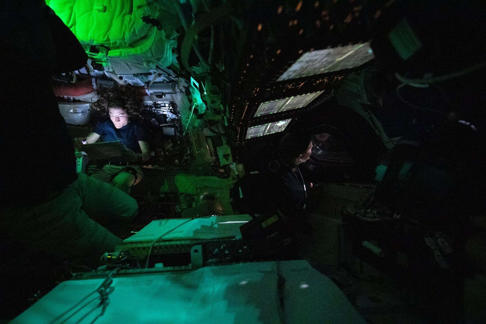 Astronaut Christina Koch is nestled into a corner of the Orion crew capsule, reading. The rest of the capsule is dark, but her face is lit by the glow from her tablet. Two of her other crew members can dimly be seen looking out the window and floating in the foreground.
