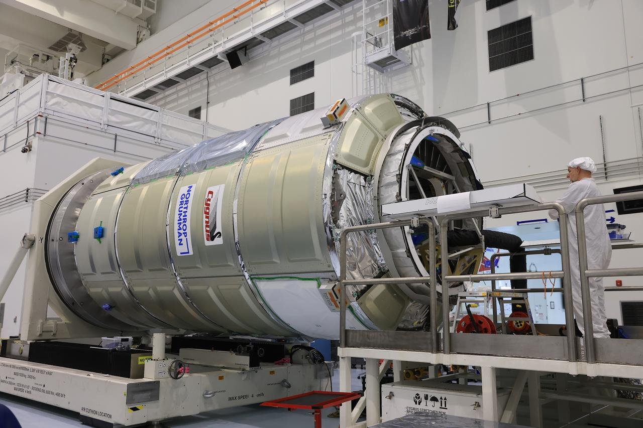 A large beige and silver cylinder-shaped capsule within a space facility is reviewed by a male technician, who is wearing a white protective suit and white head covering.