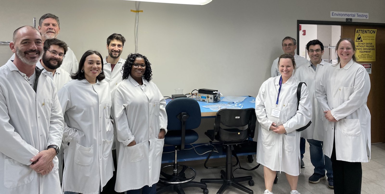 A team of 10 people pose for a photo within a laboratory. The group includes four men and two women standing on the left side, all wearing lab coats. The IVGEN Mini system is on a table in the center of the photo, and to the right are four additional people standing – two women and two men.