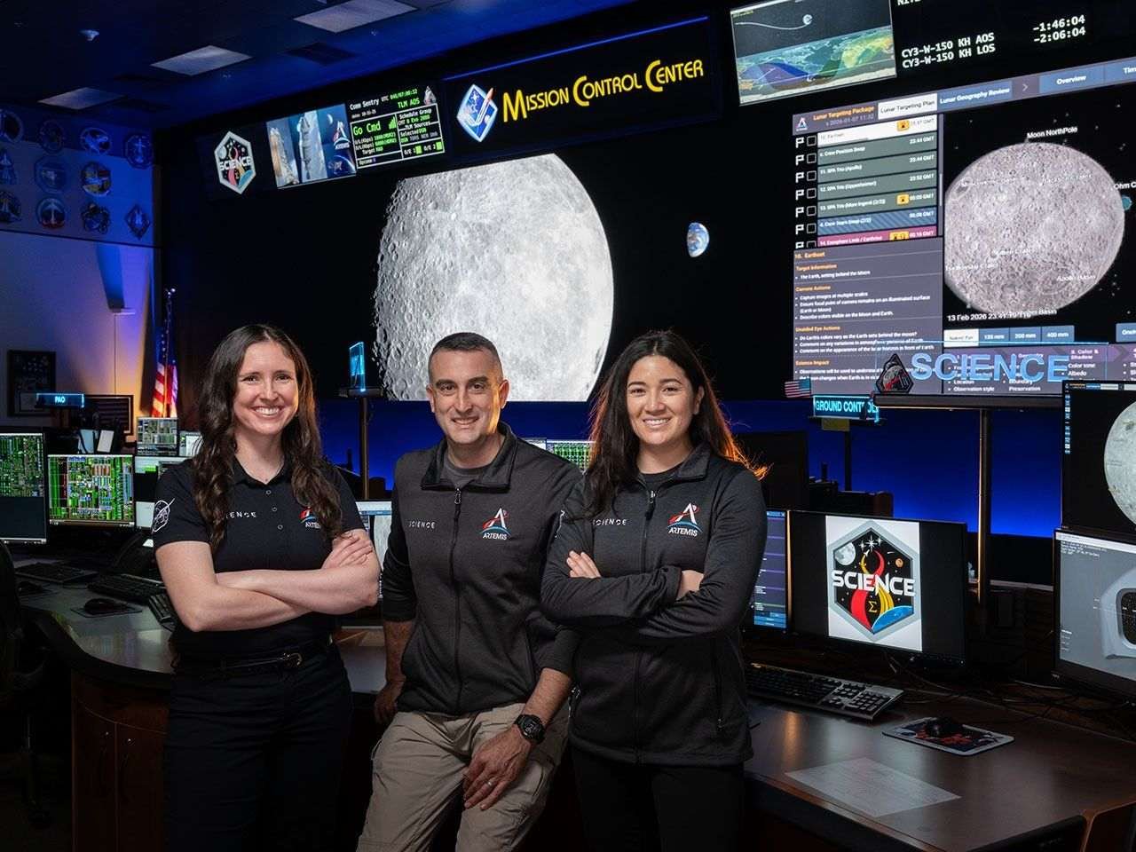 A picture of two women and a man standing at a Mission Control console desk. They are wearing shirts and jackets with the Artemis Science logo. The large display screens of Mission Control can be seen behind them, showing pictures of the Moon. The lighting in the room is dimmed and accented with blue lights.