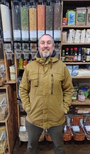 A man, in beige anorak, smiles at the camera, standing flanked by shelves filled with tidy dispensing jars and food products at point-of-sale