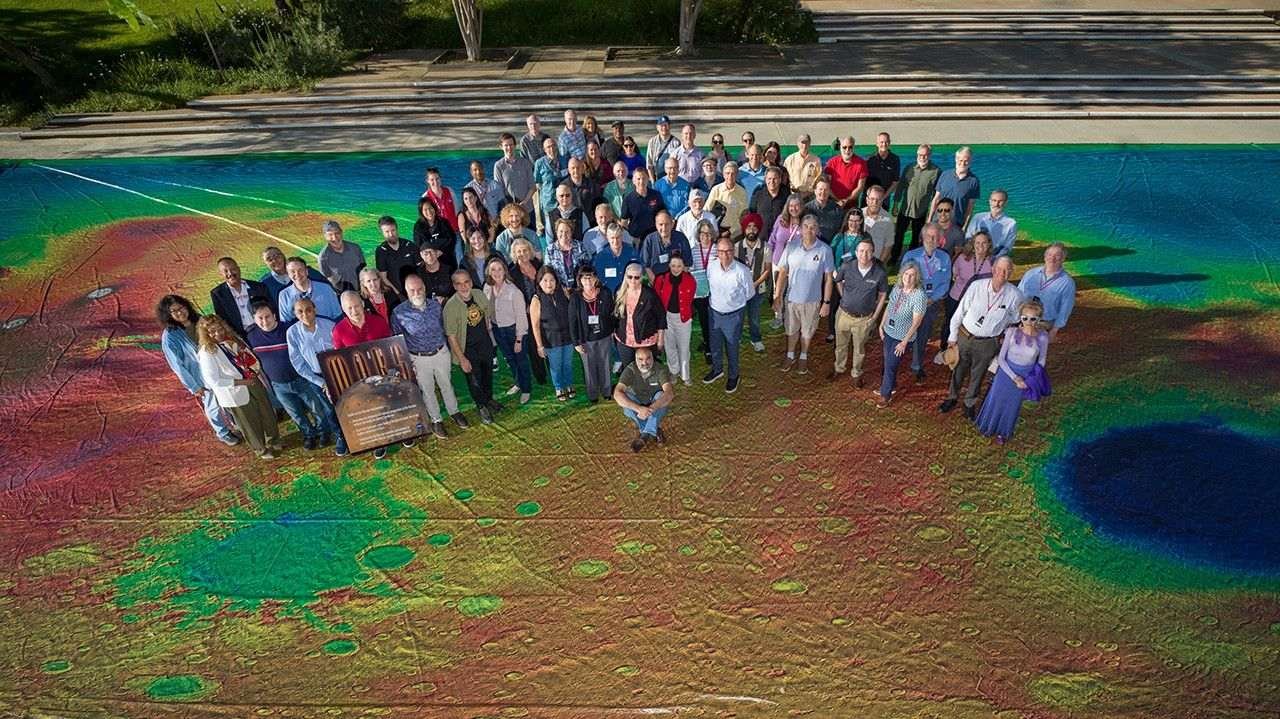 A group of several dozen scientists and engineers pose together, standing atop an auditorium-size colorful map of the Martian globe that shows patches of blue, green, red, and yellow.