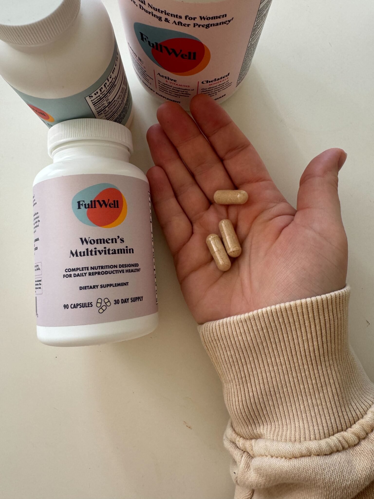 A hand holds three beige capsules next to a bottle labeled "FullWell Women's Multivitamin" and two other supplement bottles on a white surface.