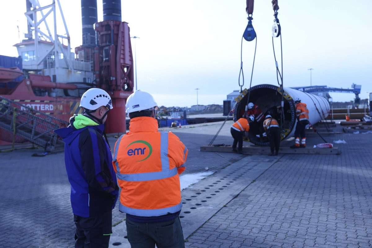 Two men in hi-vis look on as a large tubular structure - the post of a wind turbine - lies flat on an outdoor runway, with workmen and overhead hoisting equipment also within view