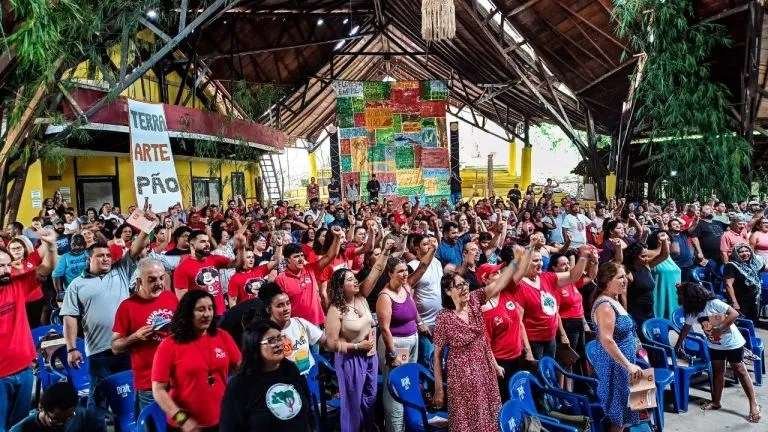 A large crown of members from the Rural Workers Movement meet in Belem at COP 30 with banners reading 'land, art, bread', their fists are raised in a celebration of solidarity. 