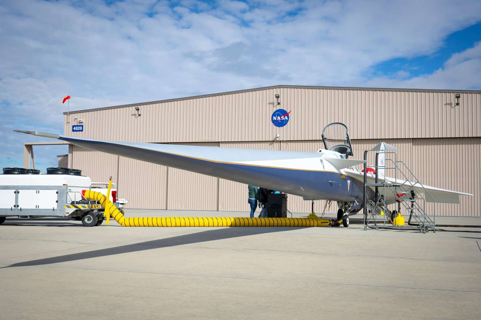 A white and blue jet airplane is parked in front of a building with large sliding doors and a NASA logo centered on the forward wall. The building is the new X-59 hangar.
