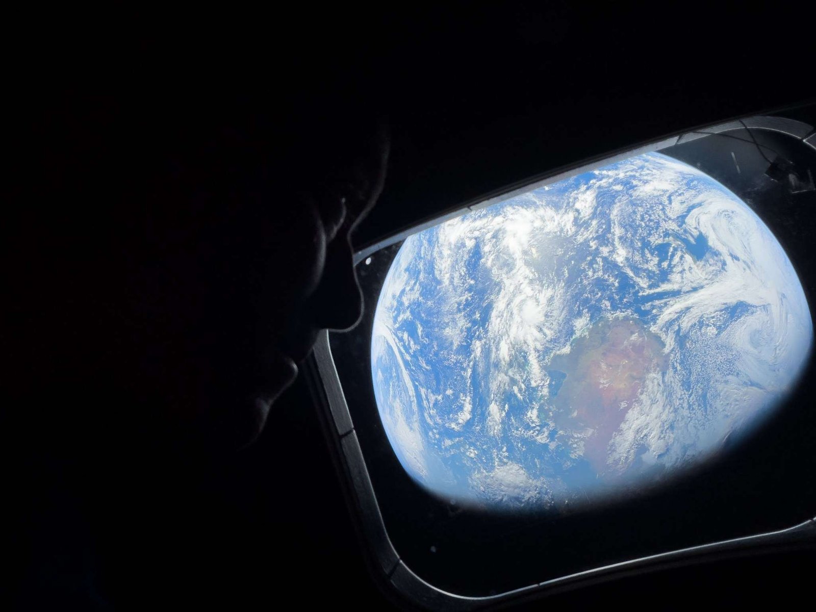 NASA astronaut Reid Wiseman looks out of the Orion spacecraft's rectangular window at Earth. He is barely visible in the dimly lit cabin, but Earth is bright. The entirety of Earth cannot be seen; in this view, Earth's round shape is cut off. Earth itself looks like a bright blue ball with swirling white clouds all over it; at center is a brown and green landmass.
