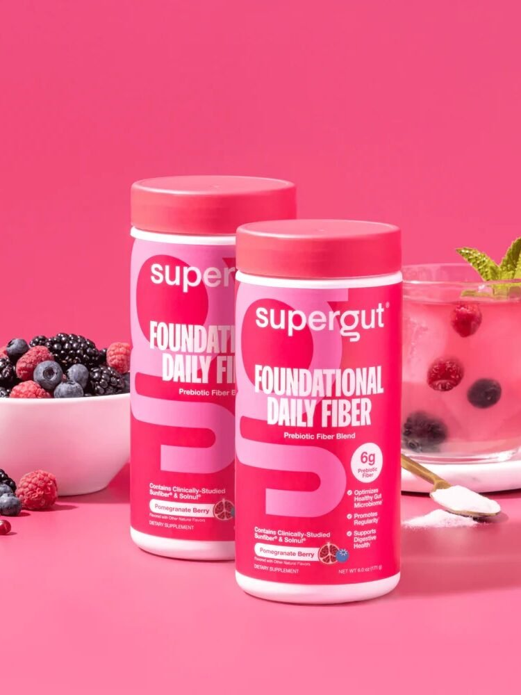 Two containers of Supergut Foundational Daily Fiber powder, a top choice in fiber supplements for women, are displayed in front of a bowl of mixed berries and a glass of pink berry drink on a pink background.