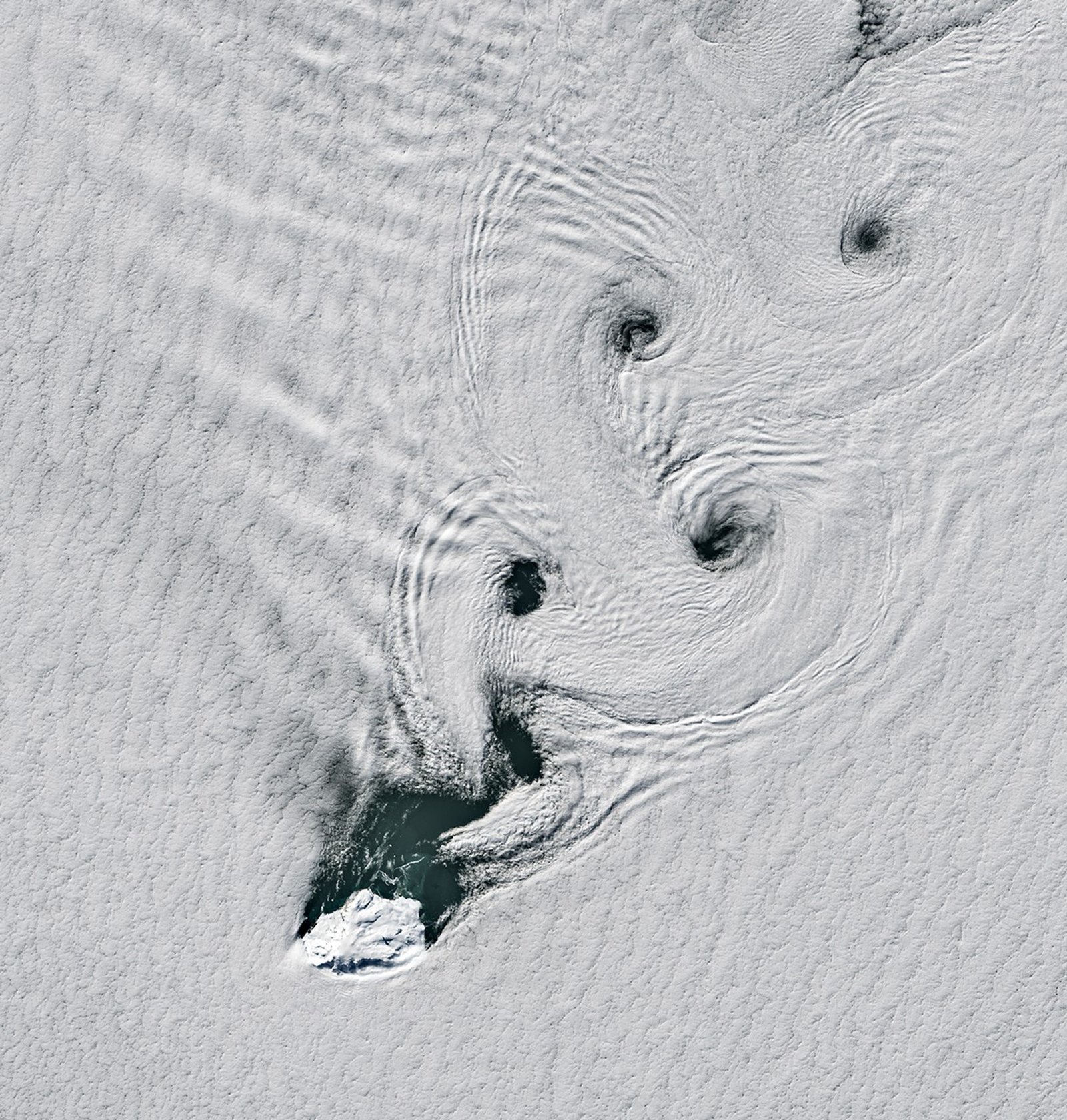 Parallel lines of cloud vortices appear downwind of a small, ice-covered island.