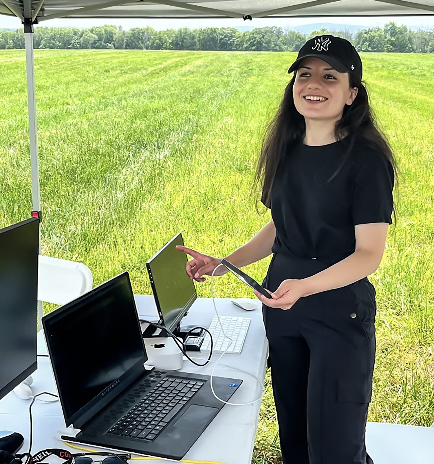 A young woman in black shirt, pants, and baseball cap stands under a tent in an open field with a table full of laptop computers used to operate a drone traffic management simulation.
