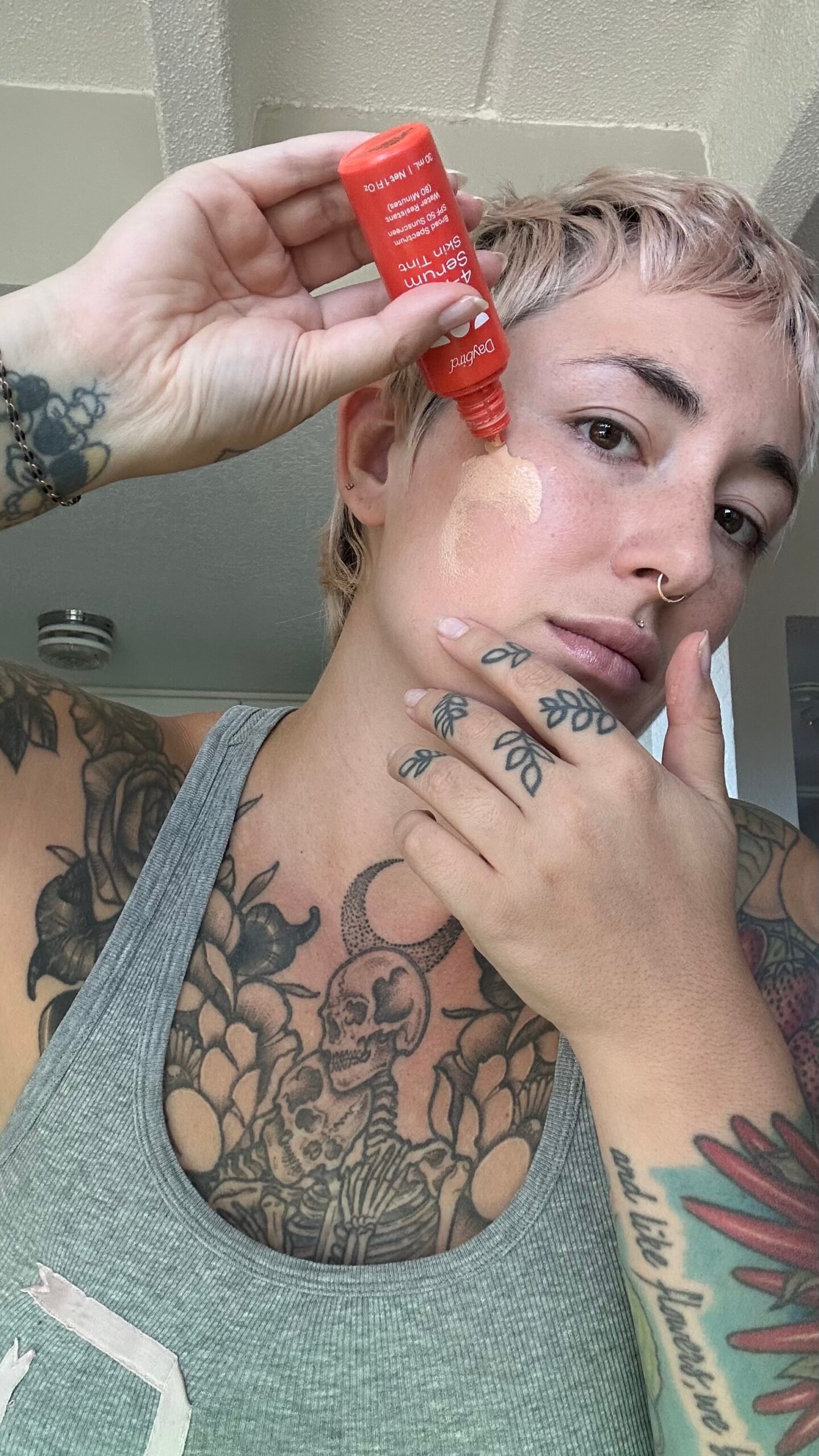 Person applying a small amount of cream or serum from a red tube onto their cheek, showing tattoos on their arm, hand, and chest, while looking at the camera.