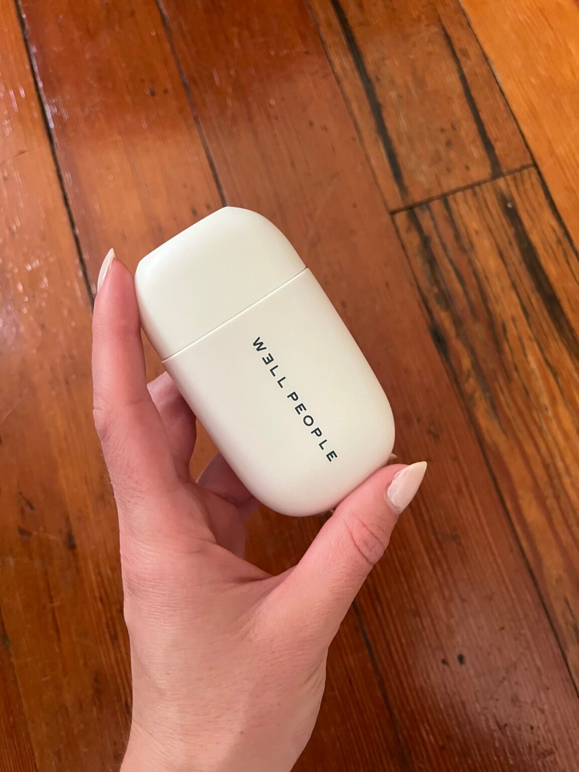 A hand holding a white, rectangular container labeled "WELL PEOPLE" against a wood floor background.