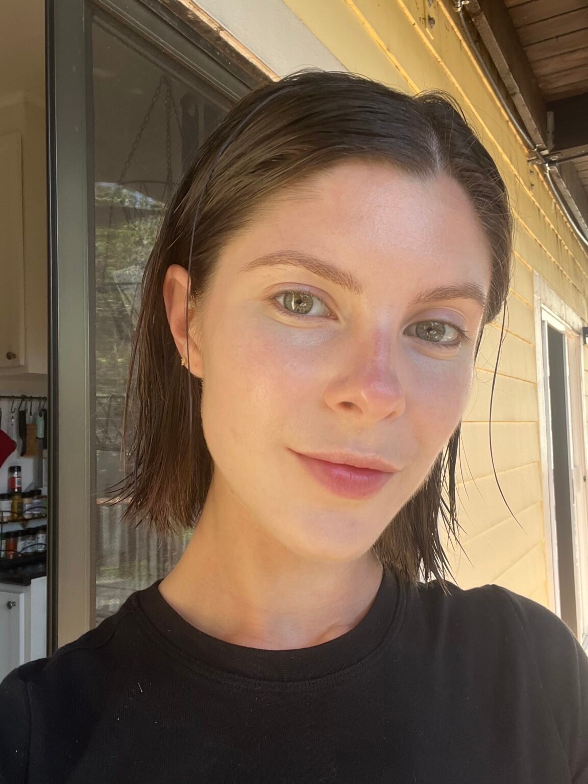 A person with short brown hair and light skin wearing a black shirt stands outdoors in natural light, close to a yellow house with a sliding glass door visible in the background.