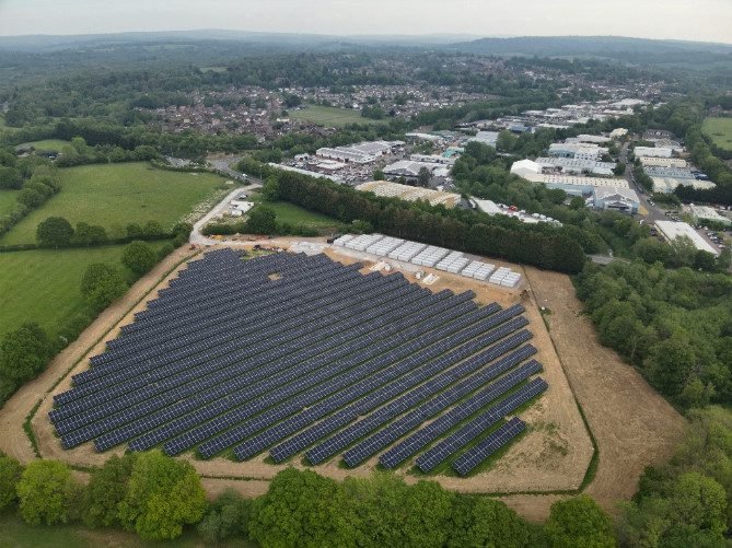 Aerial view of the Copwood VFB Energy Hub in East Sussex, showing a large ground-mounted solar array connected to rows of vanadium flow battery units, with surrounding countryside, woodland and nearby industrial buildings in the background.