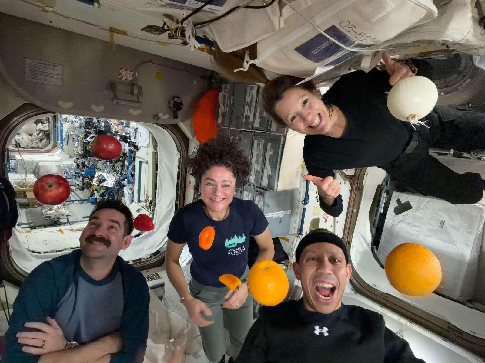 NASA astronauts Jack Hathaway (bottom left), Jessica Meir (middle left), and Chris Williams (bottom right), and ESA (European Space Agency) astronaut Sophie Adenot (top right) pose for a group photo aboard the International Space Station, as fruits and vegetables float around them.