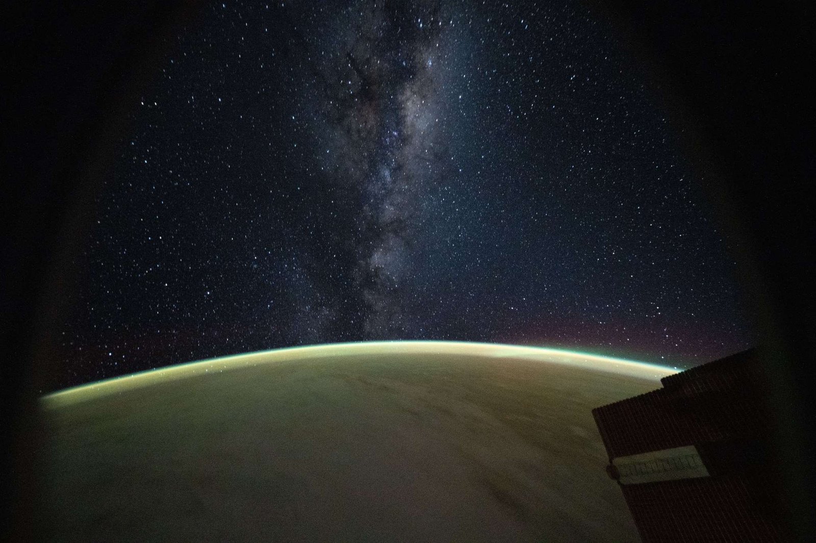 The Milky Way galaxy appears above Earth's greenish atmospheric glow. The galaxy's dust and stars can be seen against the darkness of space.