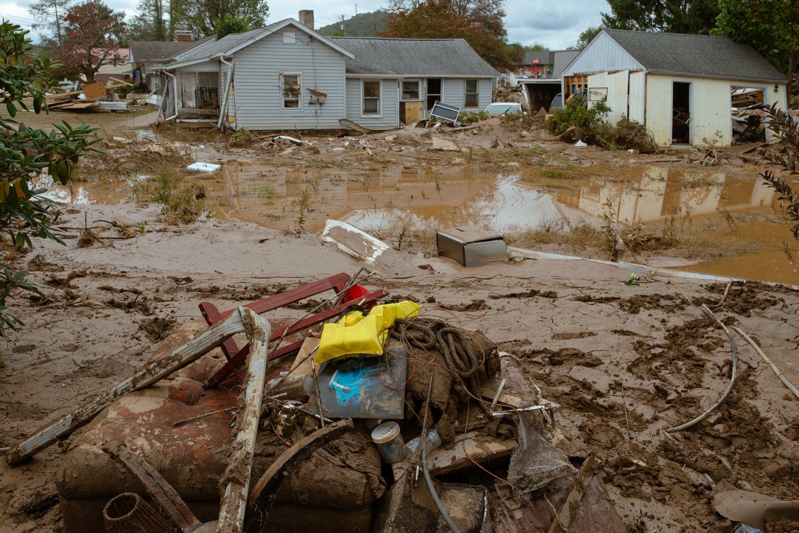 A scene of storm debris scattered across a muddy landscape. A house with major damage to the siding and windows sits in the background.