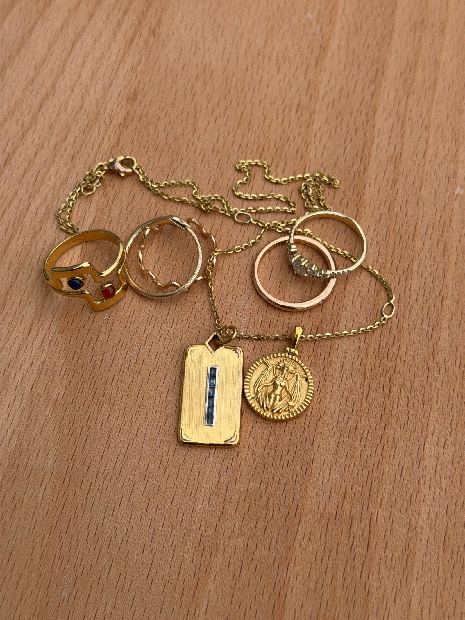 A gold chain with two pendants and four gold rings, one with colored stones, displayed on a wooden surface.