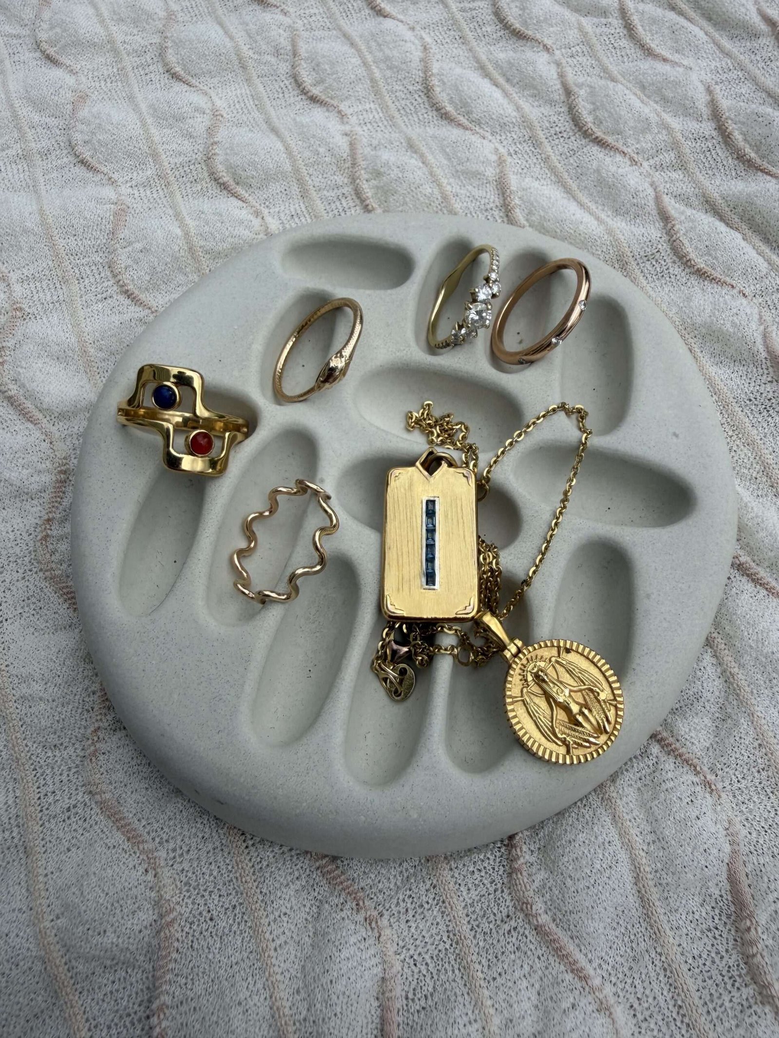 A round, gray jewelry dish displays several gold rings and two gold necklaces, one with a rectangular pendant and the other with a round medallion, on a textured fabric background.