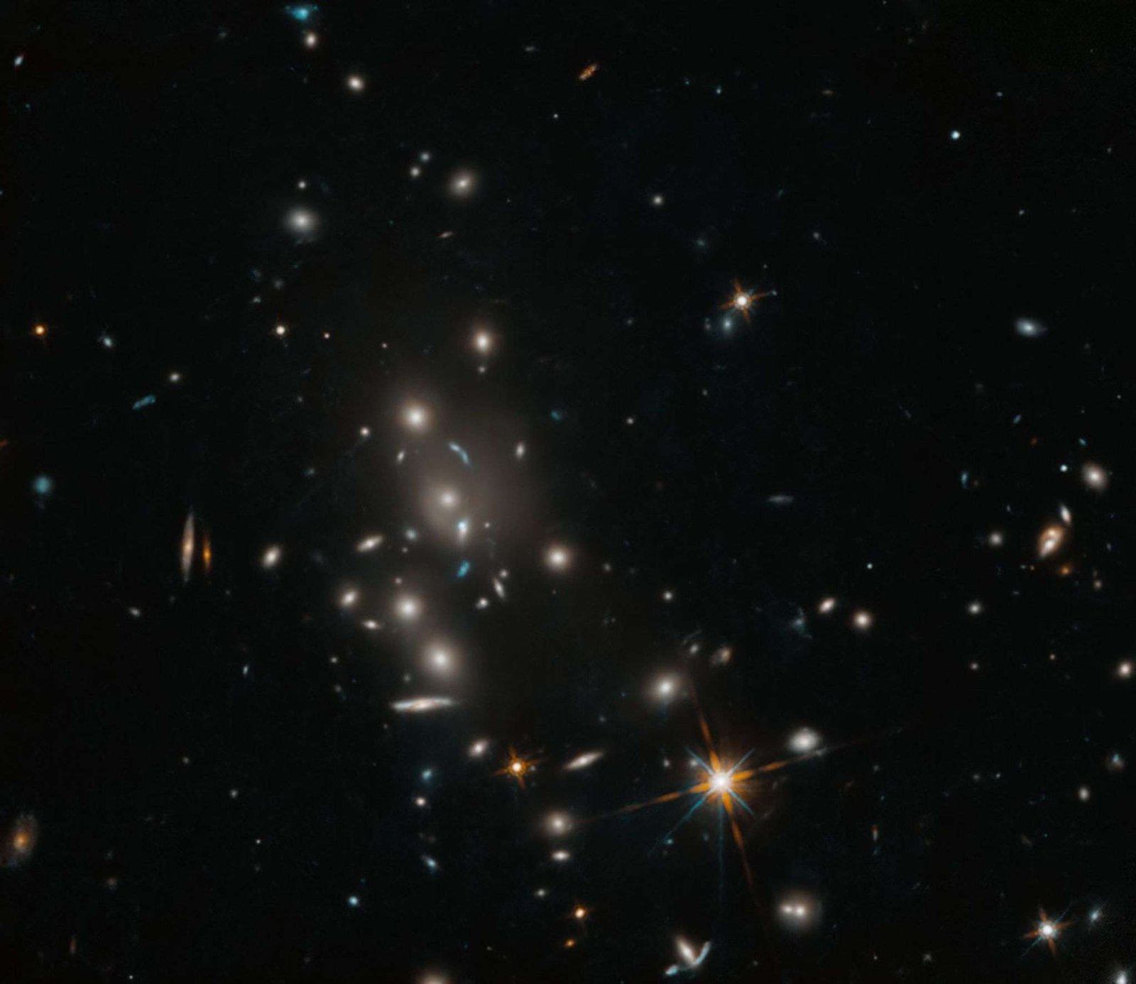 A cluster of galaxies dots the field of view. The cluster holds spiral and elliptical galaxies against a black background. A foreground star with diffraction spikes shines brightly toward the bottom, center-right of the image.