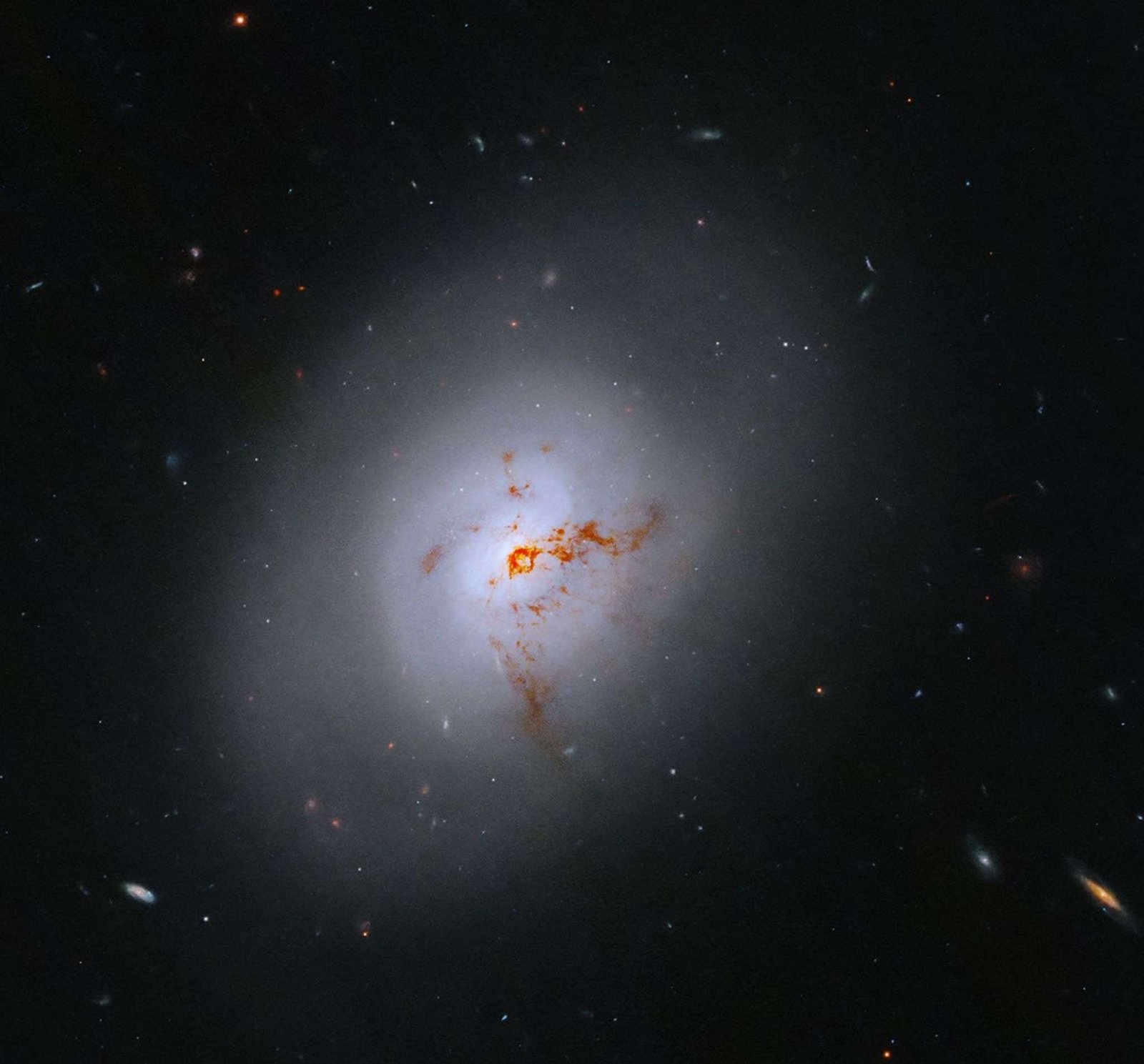 This Hubble image is of a bright lenticular galaxy (NGC 1266) seen nearly face on. Broad bright and dimmer areas of light hint at spiral structure, but there are no distinct spiral arms. Rusty-reddish-brown dust bisects the galaxy. More distant galaxies dot the black background, some even shining through the less dense regions of the bright foreground lenticular galaxy.