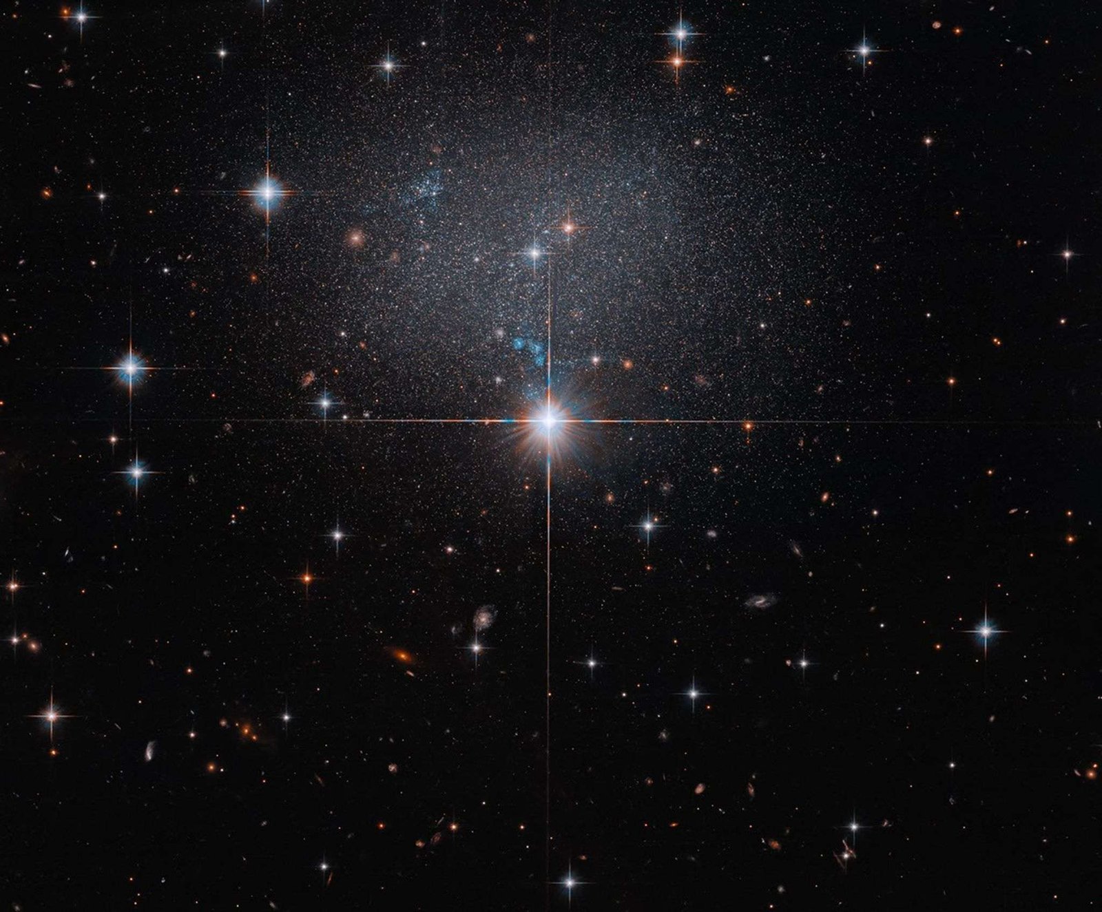 A star in the foreground shines brightly with four diffraction spikes. Above this star is a diffuse and faint dwarf irregular galaxy. The galaxy itself has bright blue and red stars.