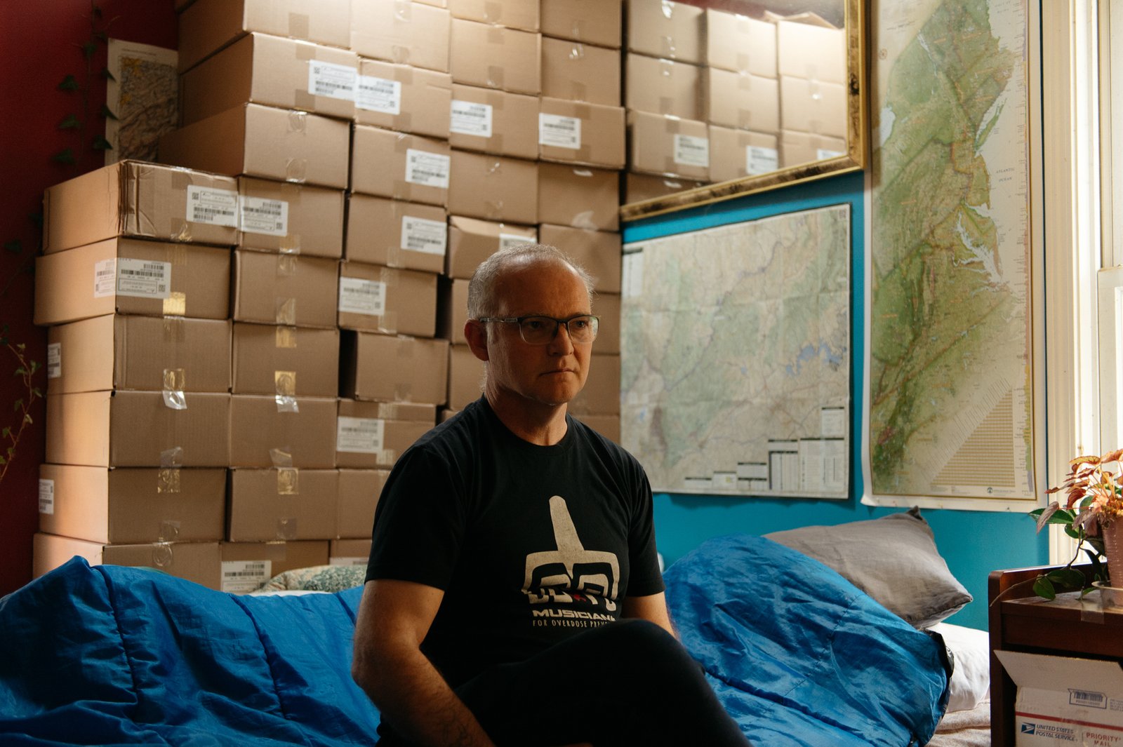 A man sits in front of a large stack of cardboard boxes containing Naloxone.