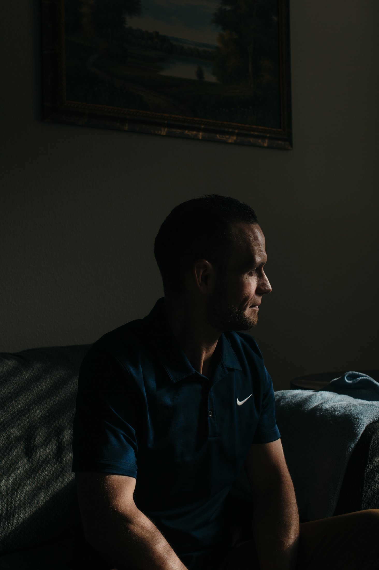A man sits in a dark room, with his face slightly illuminated by a nearby window.