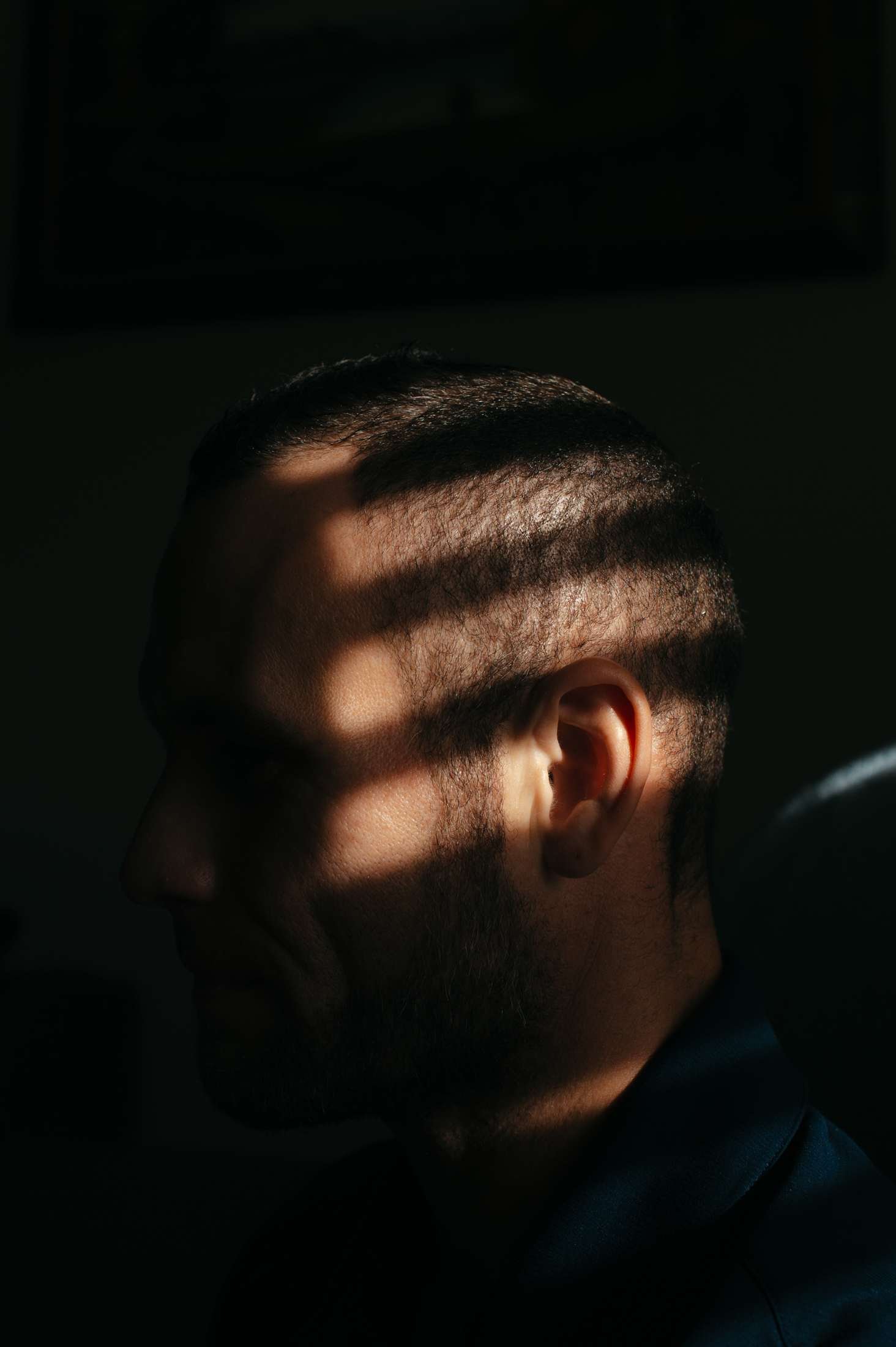 A closeup of a man's head in a dark room, with lines of light from the window blinds illuminating parts of his head.