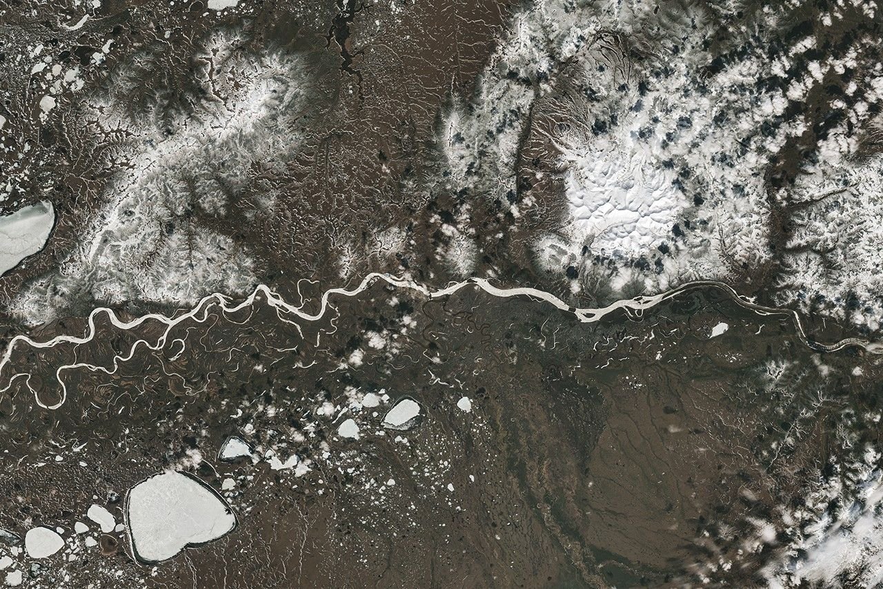 A river winds from east to west past Aniak, Alaska. Some stretches of the wide channel are still frozen over, while others contain broken-up ice. Most of the surrounding land is snow-free.