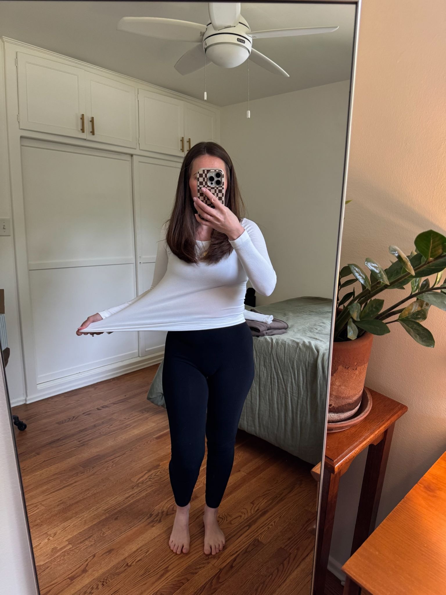 Woman standing barefoot in front of a mirror, wearing a white long-sleeve shirt and black leggings, stretching her shirt to the side in a bedroom with wooden floors and a potted plant.
