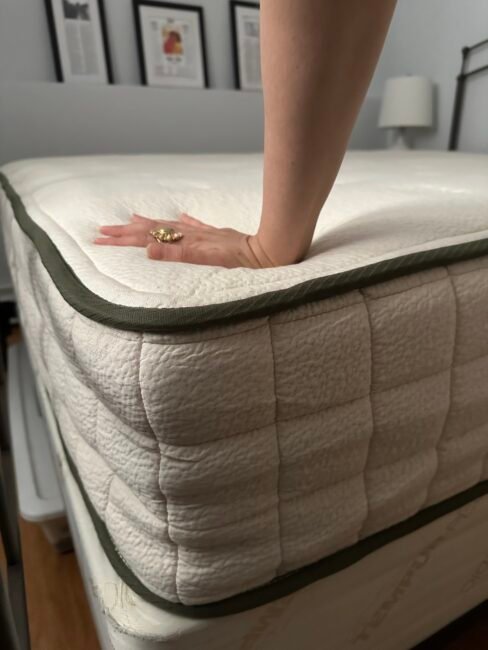 A hand presses down on the surface of a thick, white mattress with a green trim, showing its softness. Framed pictures and a lamp are visible in the background.
