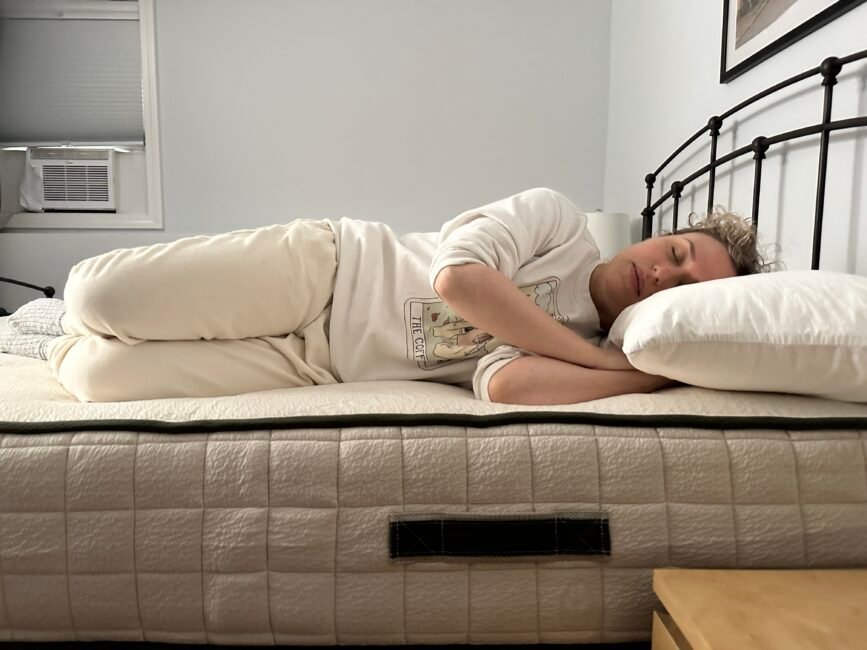 A person lies on their side on a bed, eyes closed, resting their head on a white pillow in a simply furnished bedroom.