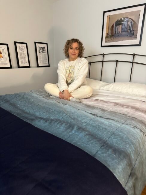 A person with curly hair sits cross-legged on a bed in a neatly decorated bedroom with framed art on the wall.