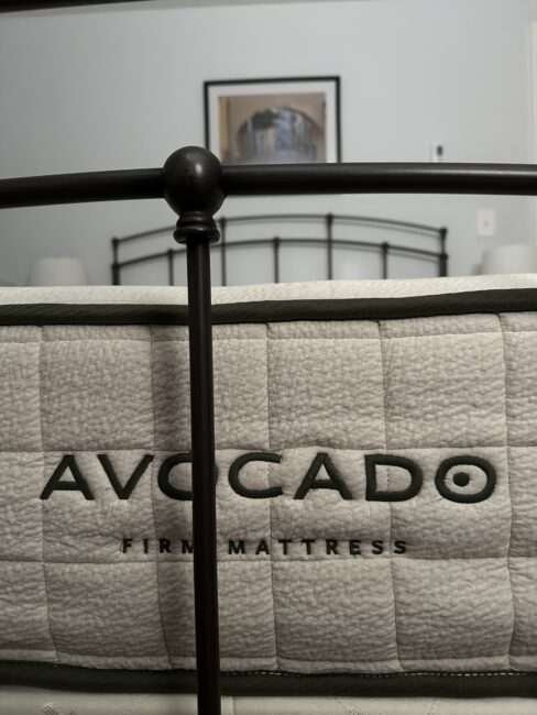 Close-up of an Avocado brand firm mattress, with a metal bed frame in the foreground and a framed picture on the wall in the background.