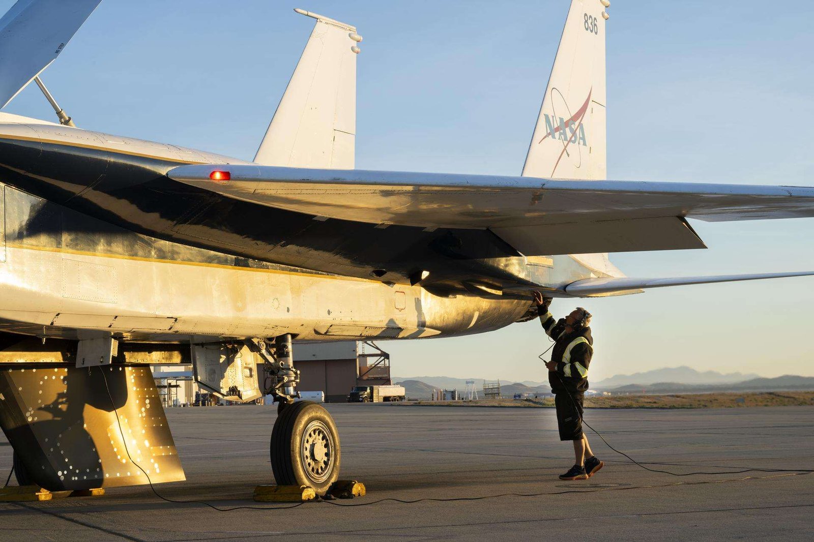 The back of a white jet aircraft’s wing and tails are seen in this image. One wheel and an attached test object, shaped like a wing, hangs from the bottom of the jet. A man in a black jacket, with yellow reflective coating, stands underneath and raises one hand to the aircraft. The man is wearing ear protection. Desert sand and mountains are seen in the background.
