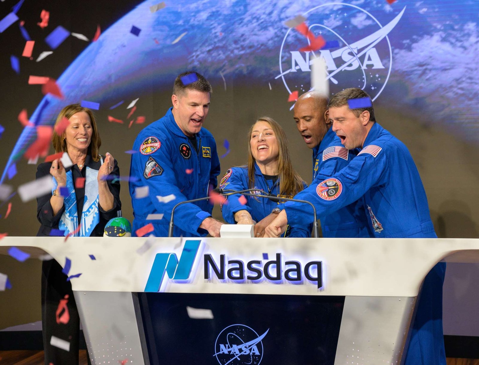 Nasdaq Chair and Chief Executive Officer Adena T. Friedman, left, and NASA’s Artemis II crewmembers CSA (Canadian Space Agency) astronaut Jeremy Hansen, and NASA astronauts Christina Koch, Victor Glover, and Reid Wiseman, right, ring the closing bell of the Nasdaq market session. They're standing behind a wide white podium with the Nasdaq logo and the word "Nasdaq." There is a screen on the front of the podium displaying the NASA meatball logo. Friedman is standing a little further away from the podium. Red, white, and blue confetti falls from the air on the left side of the image.