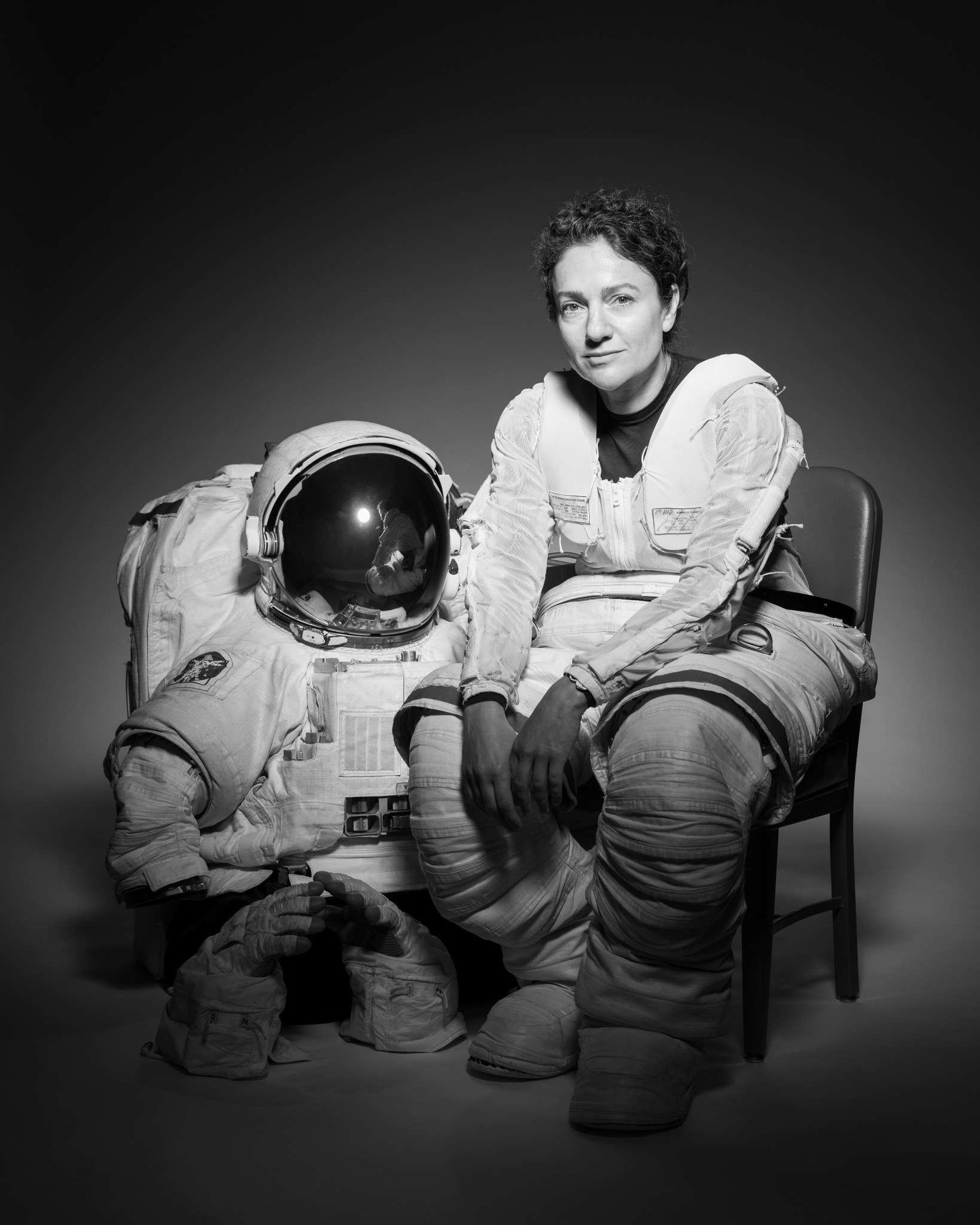 NASA Astronaut Jessica Meir sits in a chair. Her body is slightly turned to the right as she looks forward. She is wearing the bottom half of a spacesuit; the top half and gloves are on the floor next to the chair.