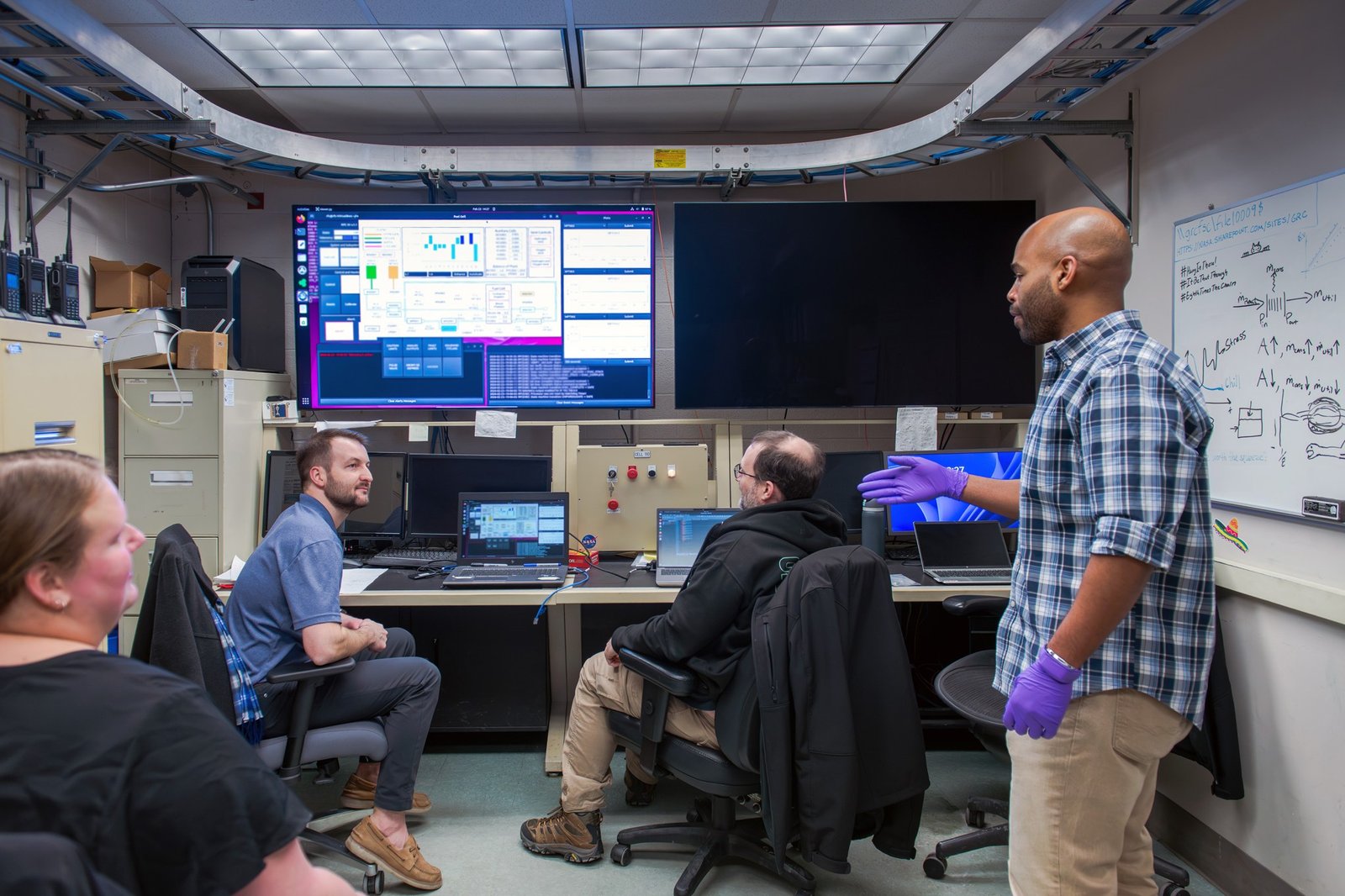Two researchers stand in a control room and converse with two other researchers sitting in chairs behind colorful monitors and laptops showing testing data. A whiteboard with equations is shown to their right.
