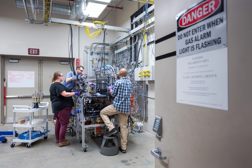 Three researchers reach inside a system filled with tubes, wires, and hardware inside a laboratory. A fuel cell sits on a white multi-tiered wheeled cart to their left along with boxes, wires, and other equipment. In the foreground is a open door with a sign that says "Caution: do not enter when gas alarm is flashing" along with another sign giving information about the laboratory. A written-on whiteboard can be seen hanging on the double doors behind the group.