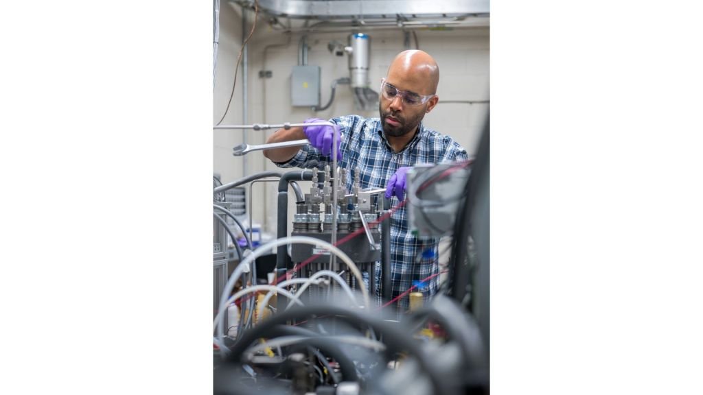 A researcher wearing purple gloves, safety glasses, and a blue plaid shirt reaches uses wrenches to adjust components inside a system that is filled with wires and hardware.