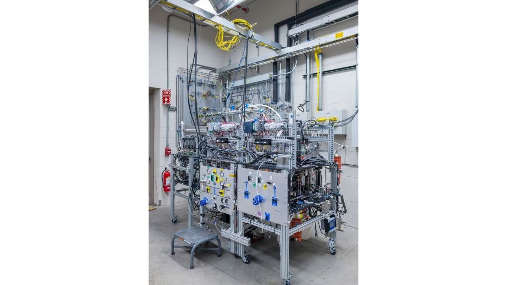 Numerous tubes, wires, and sensors are connected to a large grey system and the wall of a laboratory. A stepstool is visible to the left of the technology, and a fire extinguisher and fire alarm system are seen behind it.