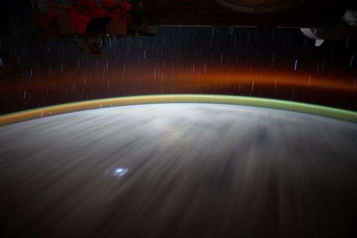 A time-lapse image of Earth from space shows two layers of airglow above Earth's surface: a band of green near the surface and a band of red higher up, with a gap of dark space in between. Star trails are visible in the background above Earth. On Earth, clouds appear as white streaks, with a flash of lightning near the bottom.